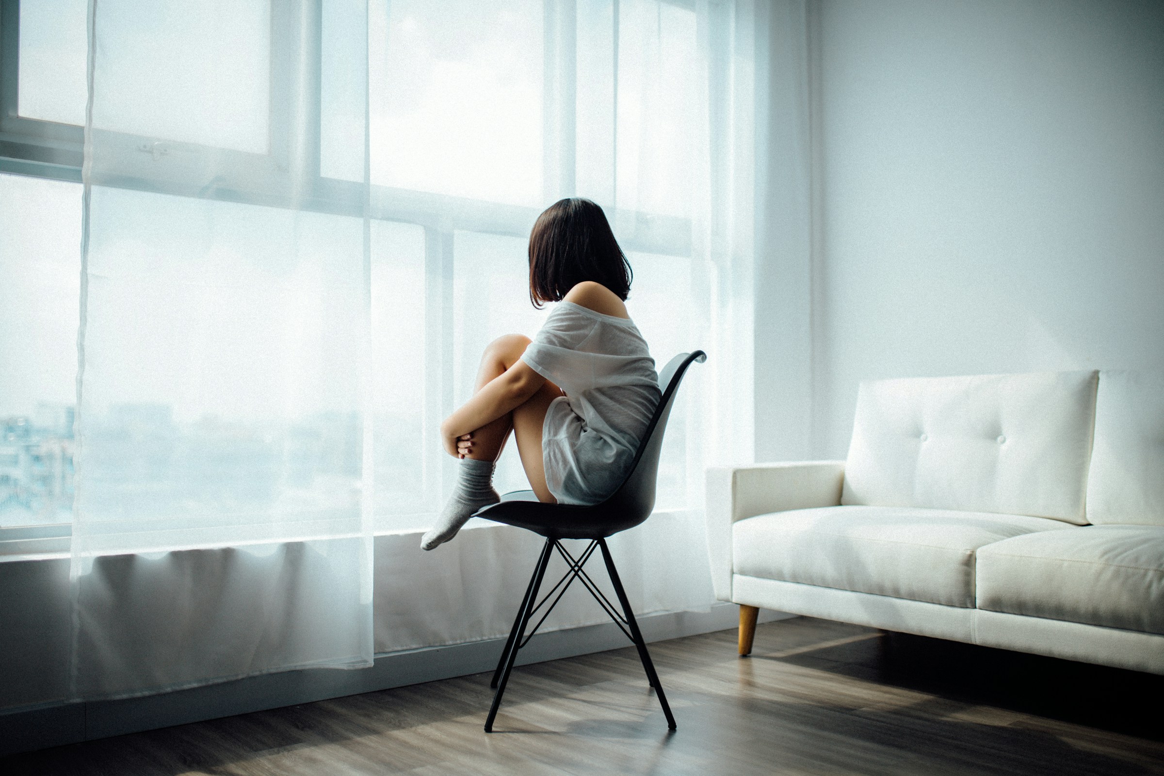 Woman sitting alone on a chair by a large bright window, looking out